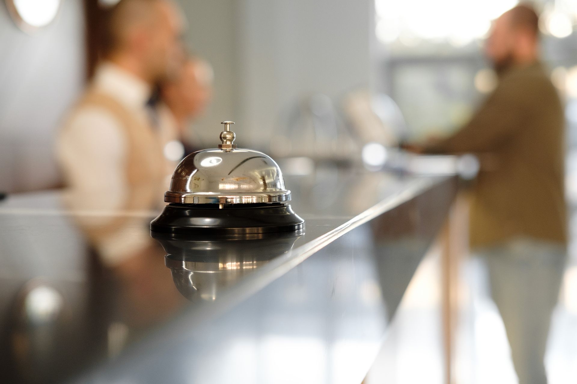 Silver bell on hotel reception desk, blurred staff and guest in background.