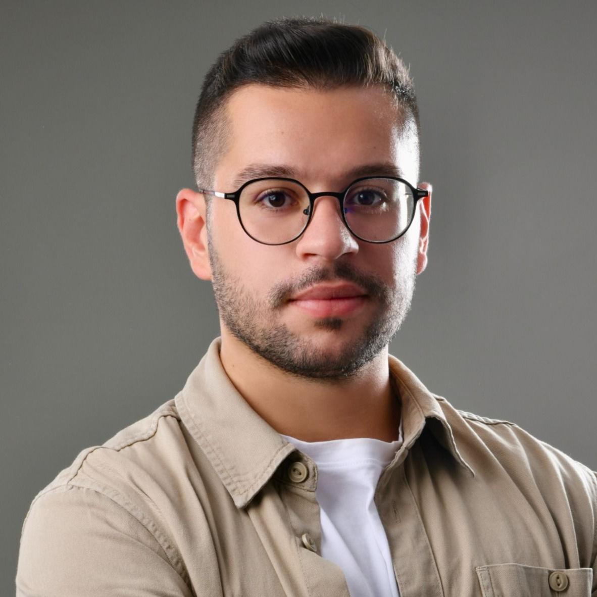 Man with dark hair, glasses, and a tan shirt, looking at the camera against a grey background.