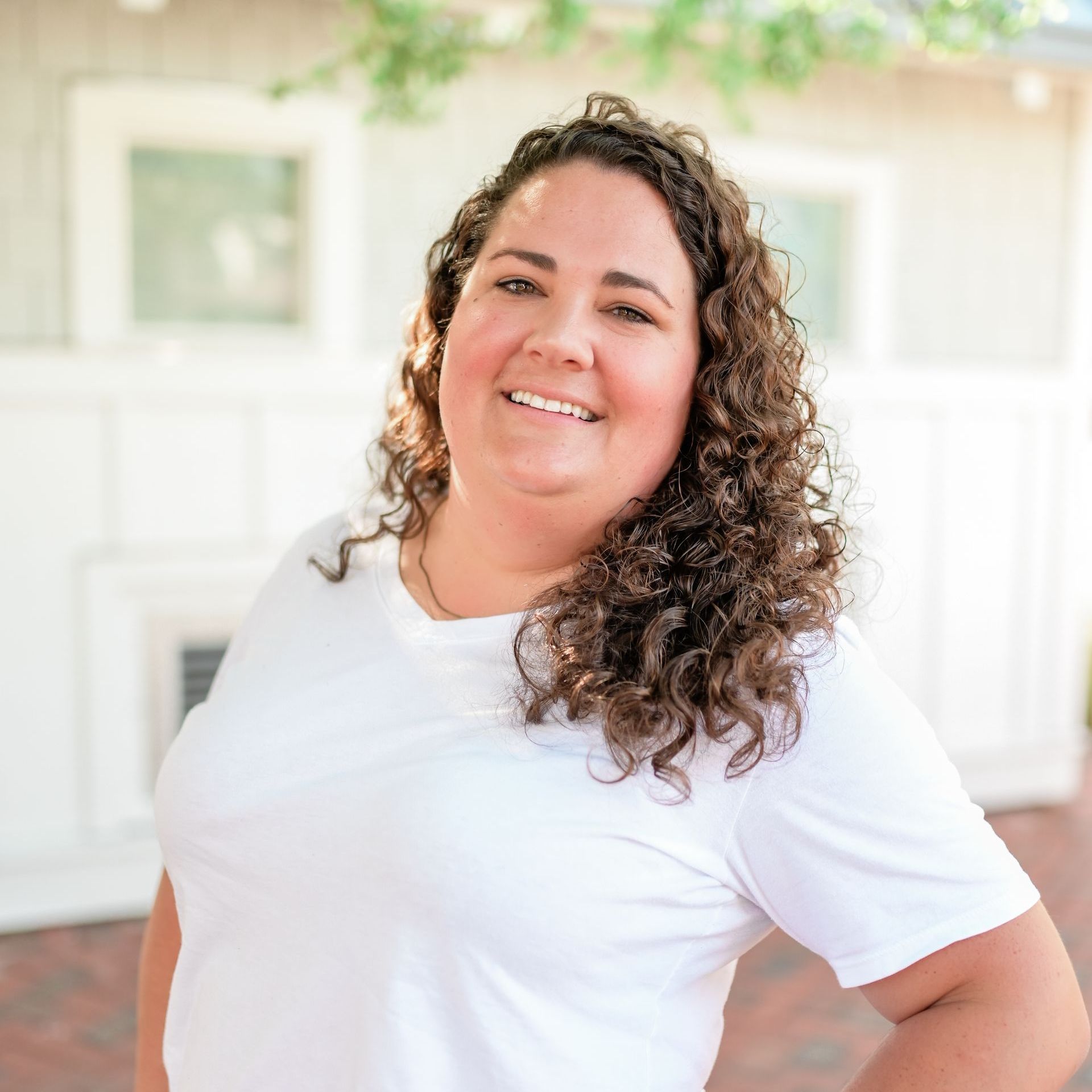 A woman with curly hair is wearing a white t-shirt and smiling.