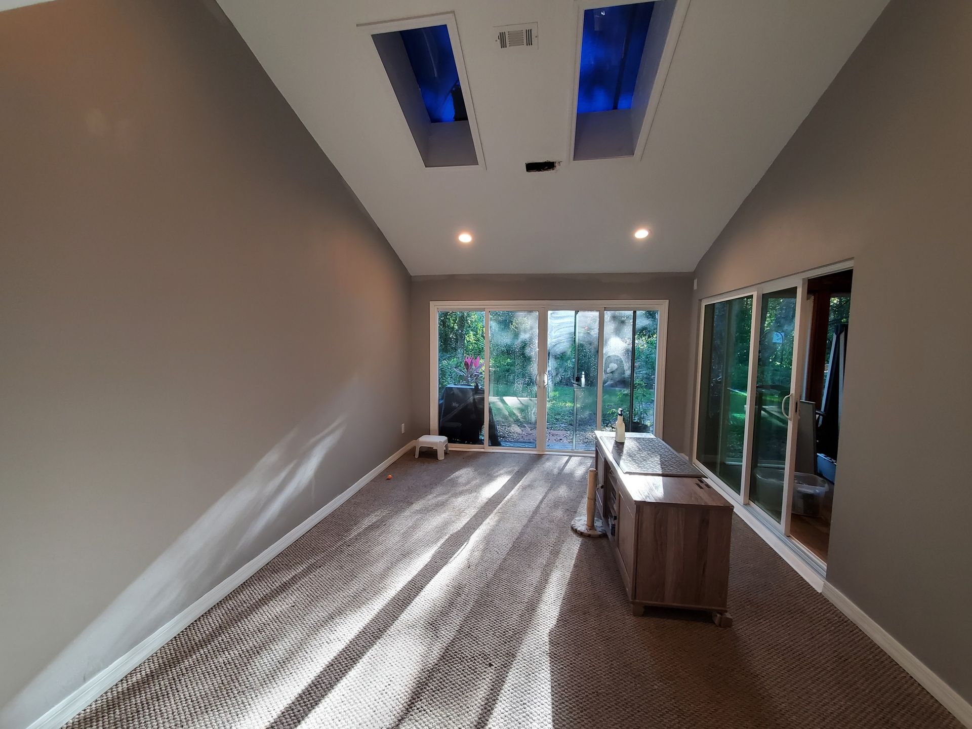 Room with tan walls, angled ceiling, skylights, and sliding glass doors. Brown carpet. Sunlight streams in.
