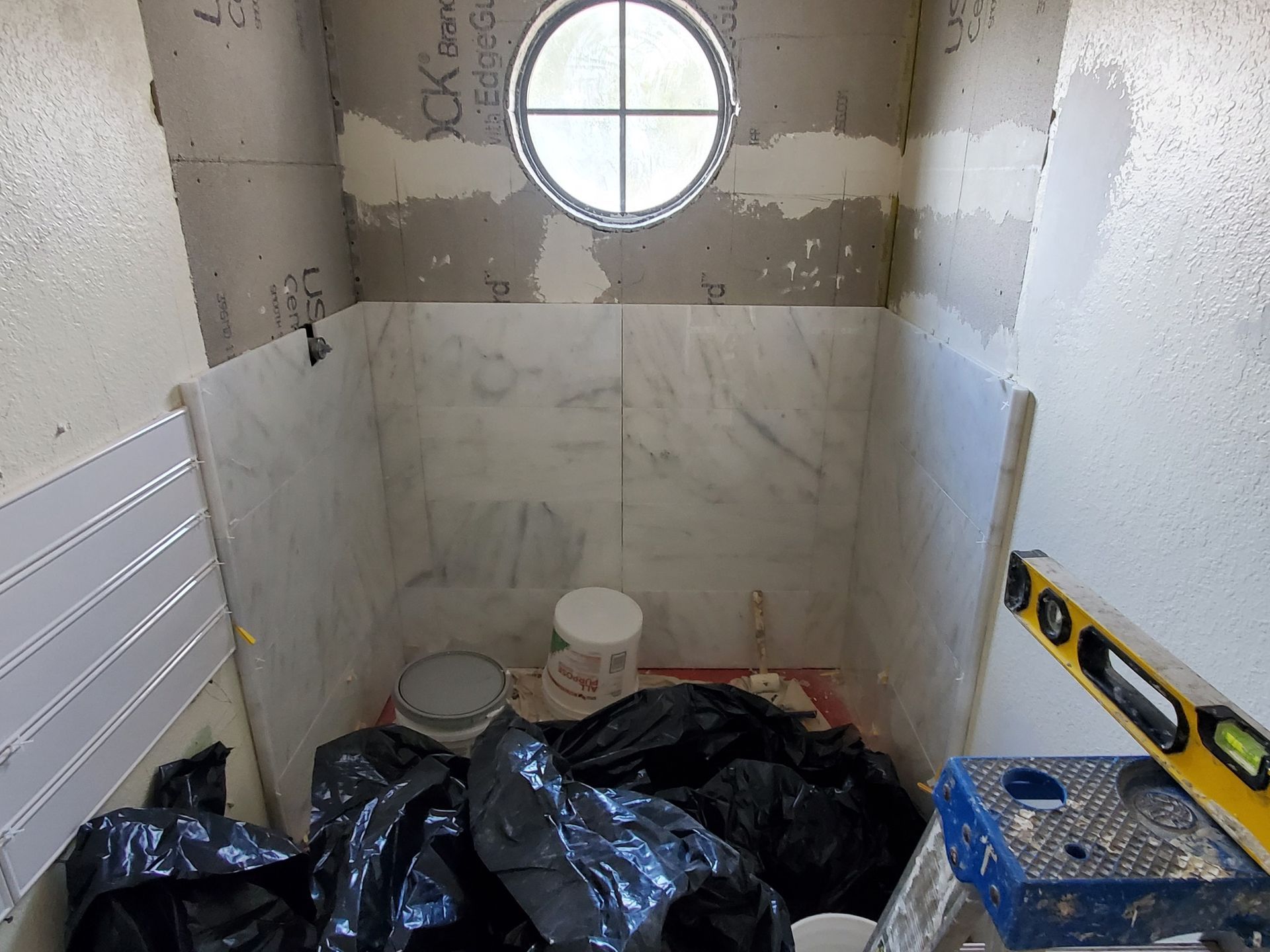 Bathroom under construction: marble tile on walls, round window, black trash bags, tools, and materials.
