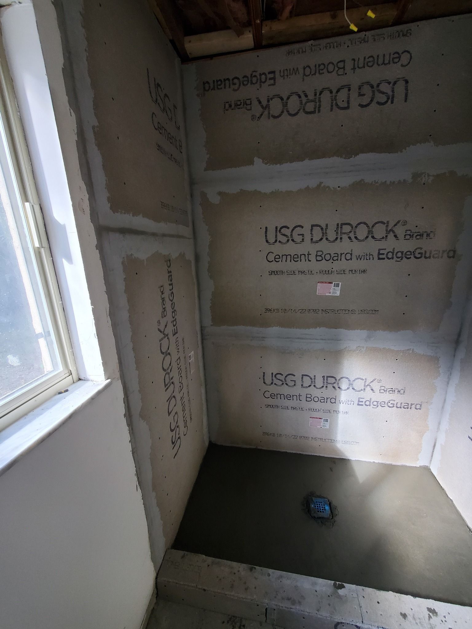 Shower stall under construction, cement board walls, concrete floor, drain visible, unfinished, near a window.