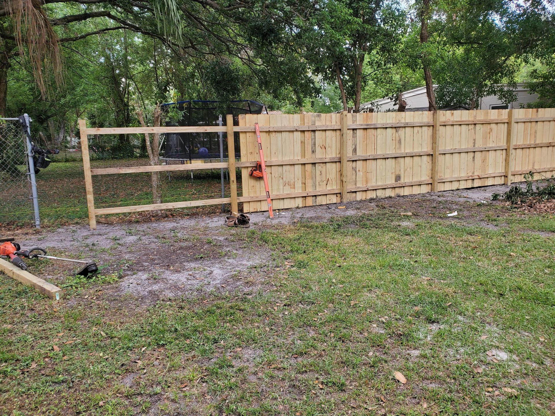 A new wooden fence built alongside a chain-link fence in a grassy yard, construction tools nearby.