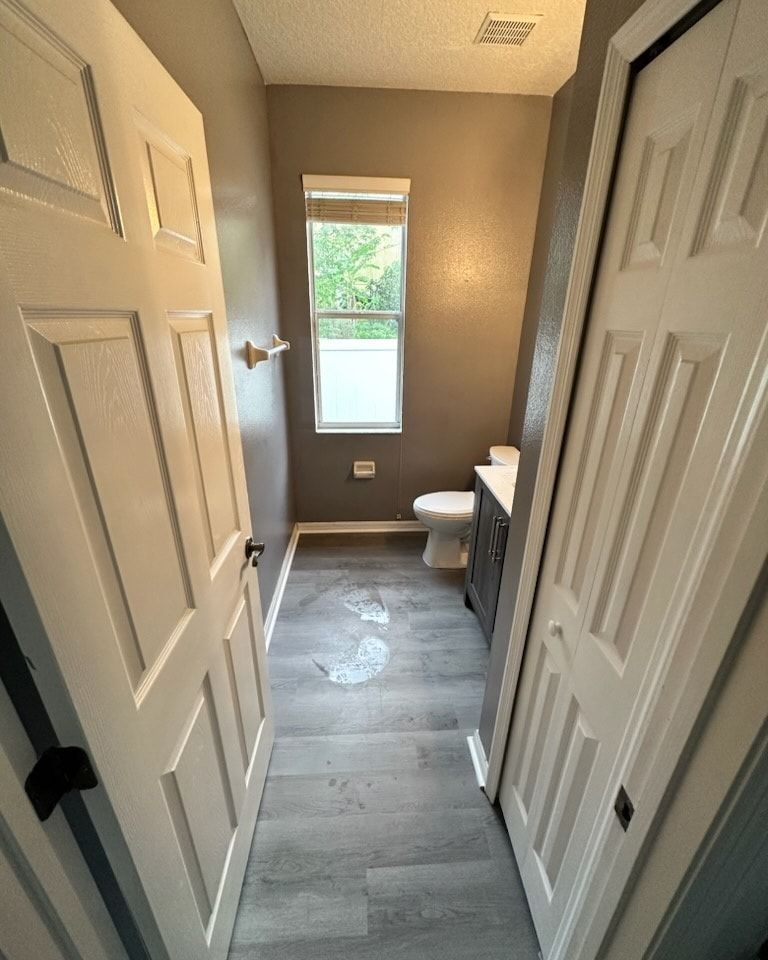 Narrow bathroom with gray walls, white doors, a toilet, window, and wood-look floor.