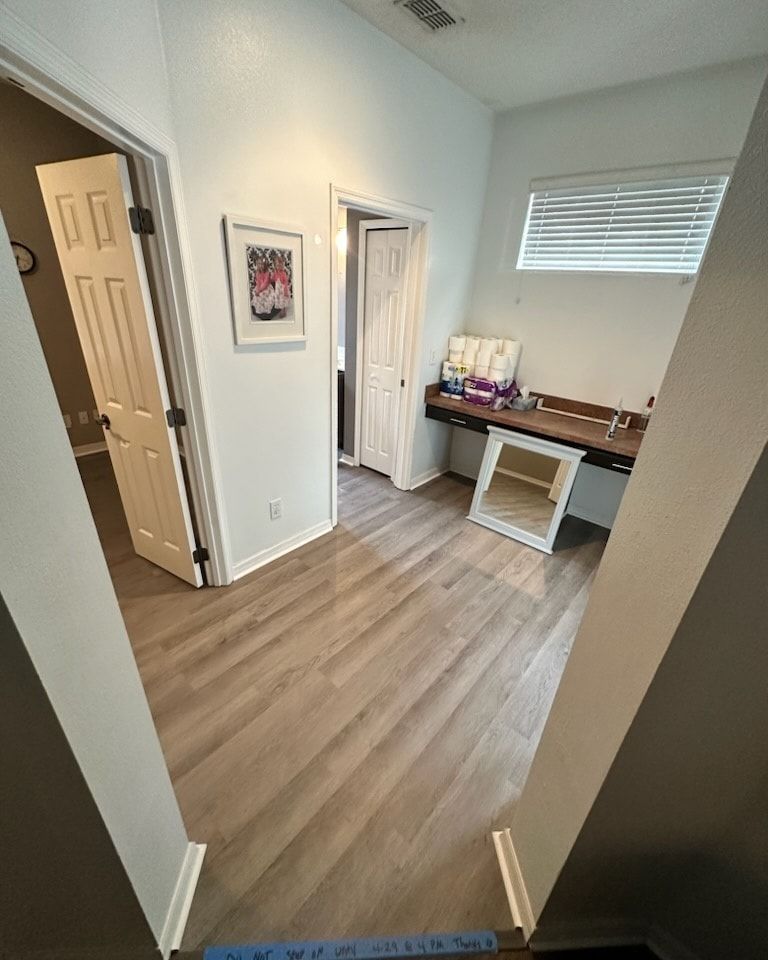 Hallway with wood-look floors, white walls, and three open doors leading to rooms. A desk with a mirror and window are visible.