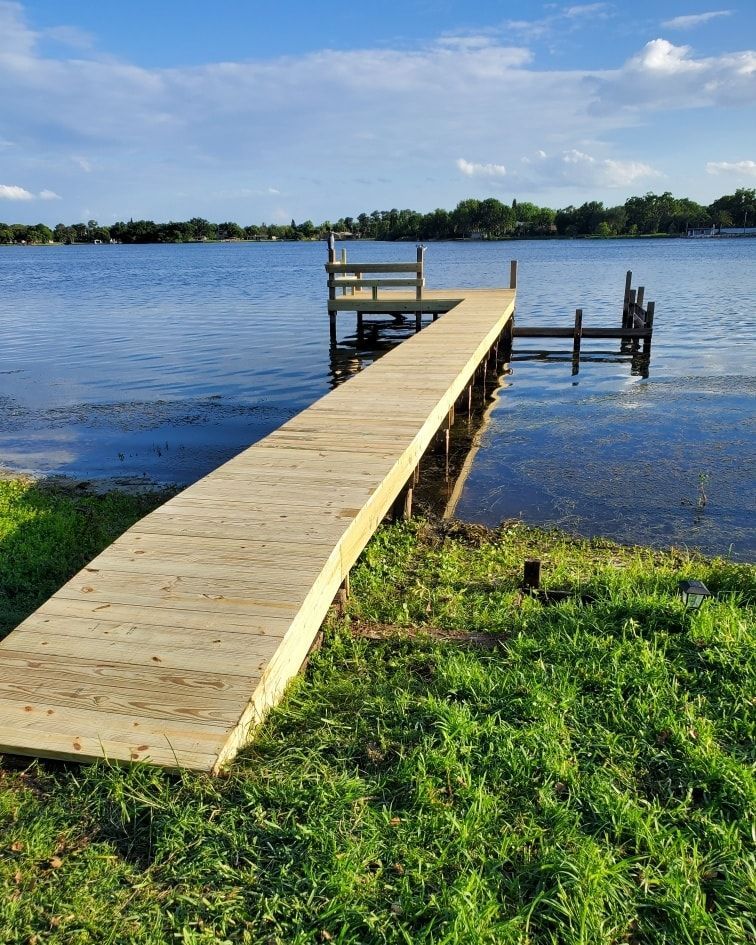 Wooden dock extends into a calm lake with a grassy shoreline and blue sky.