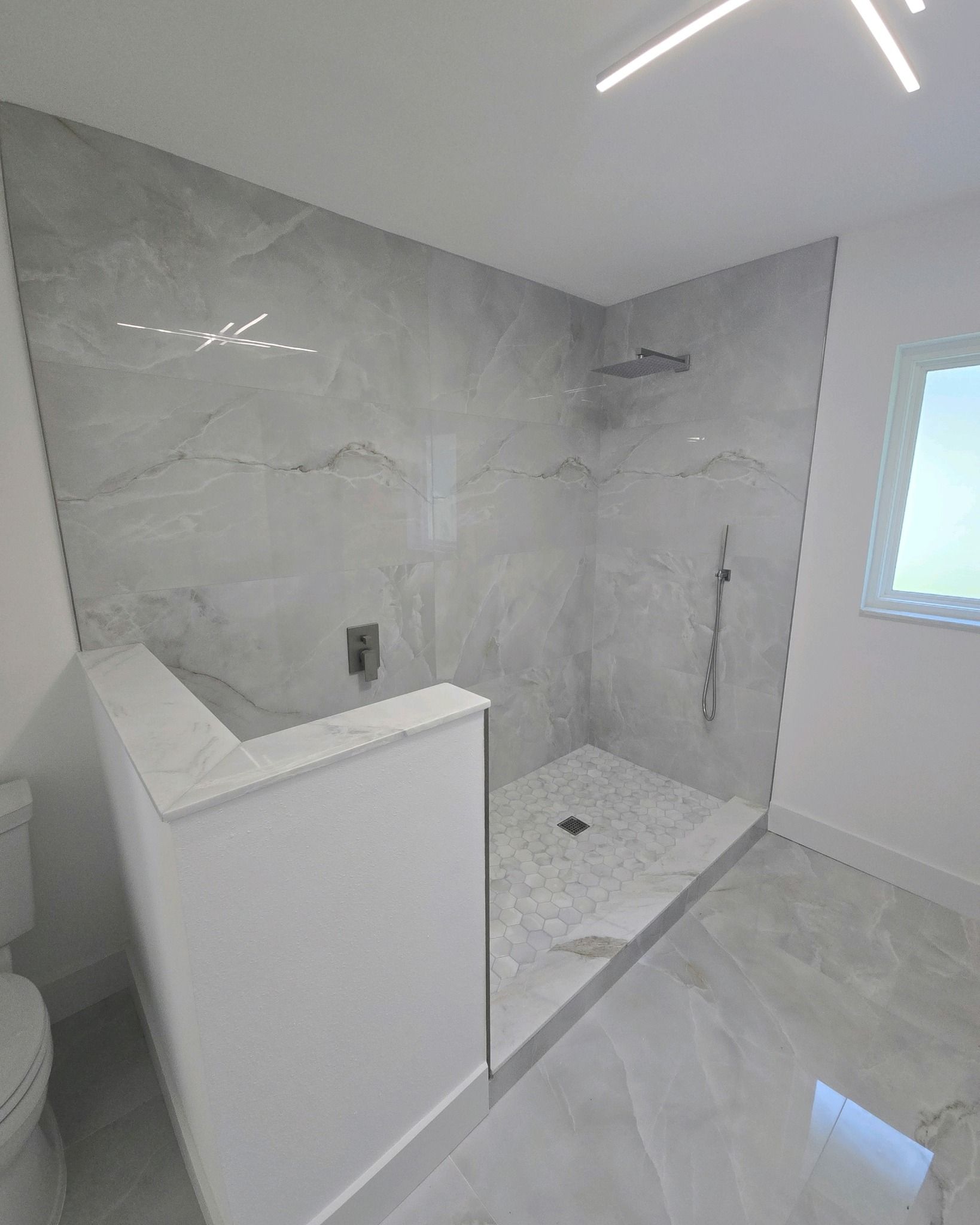 Modern bathroom with gray tiled shower and marble accents, with a window on the right.