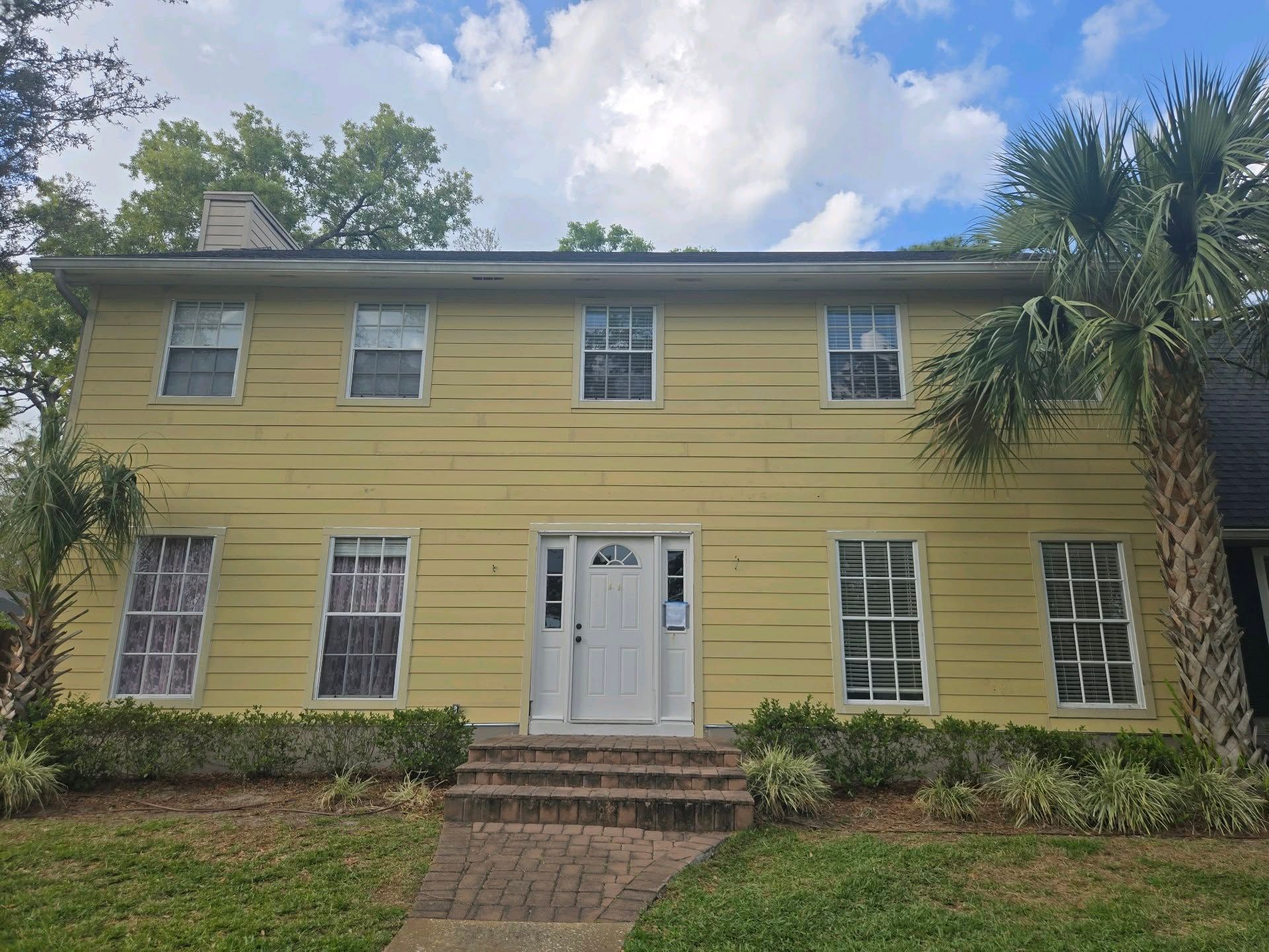Two-story yellow house with white trim. Palm trees frame the exterior. Blue sky with clouds.