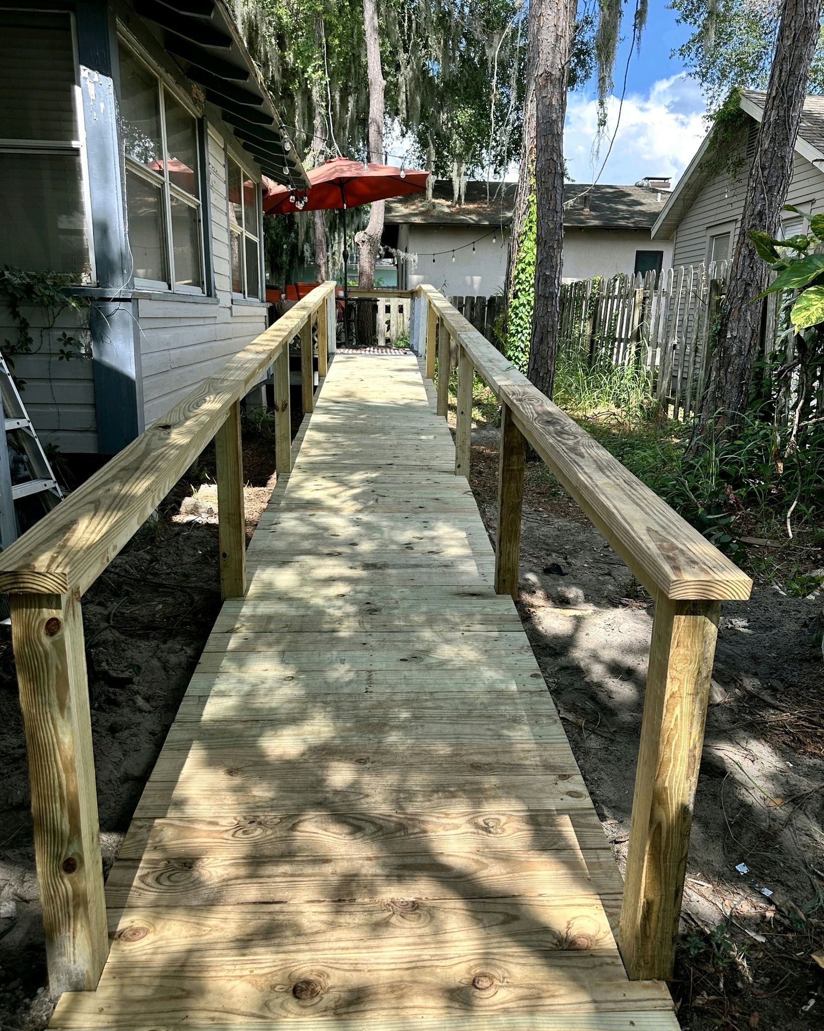 Wooden ramp with handrails leading to a building. Outdoor setting with trees and a red umbrella.