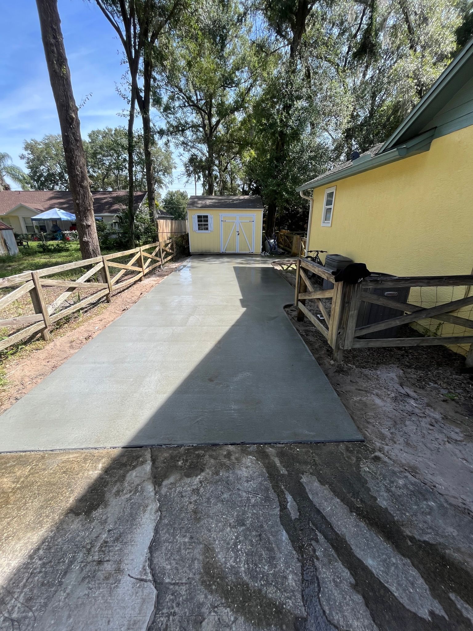 A concrete driveway leading to a yellow house with a wooden fence.