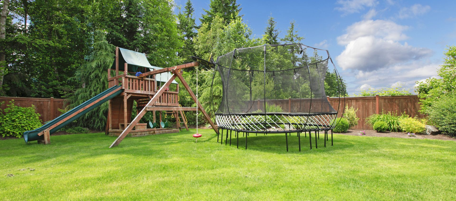 Green backyard with wooden playset and trampoline under a blue sky.