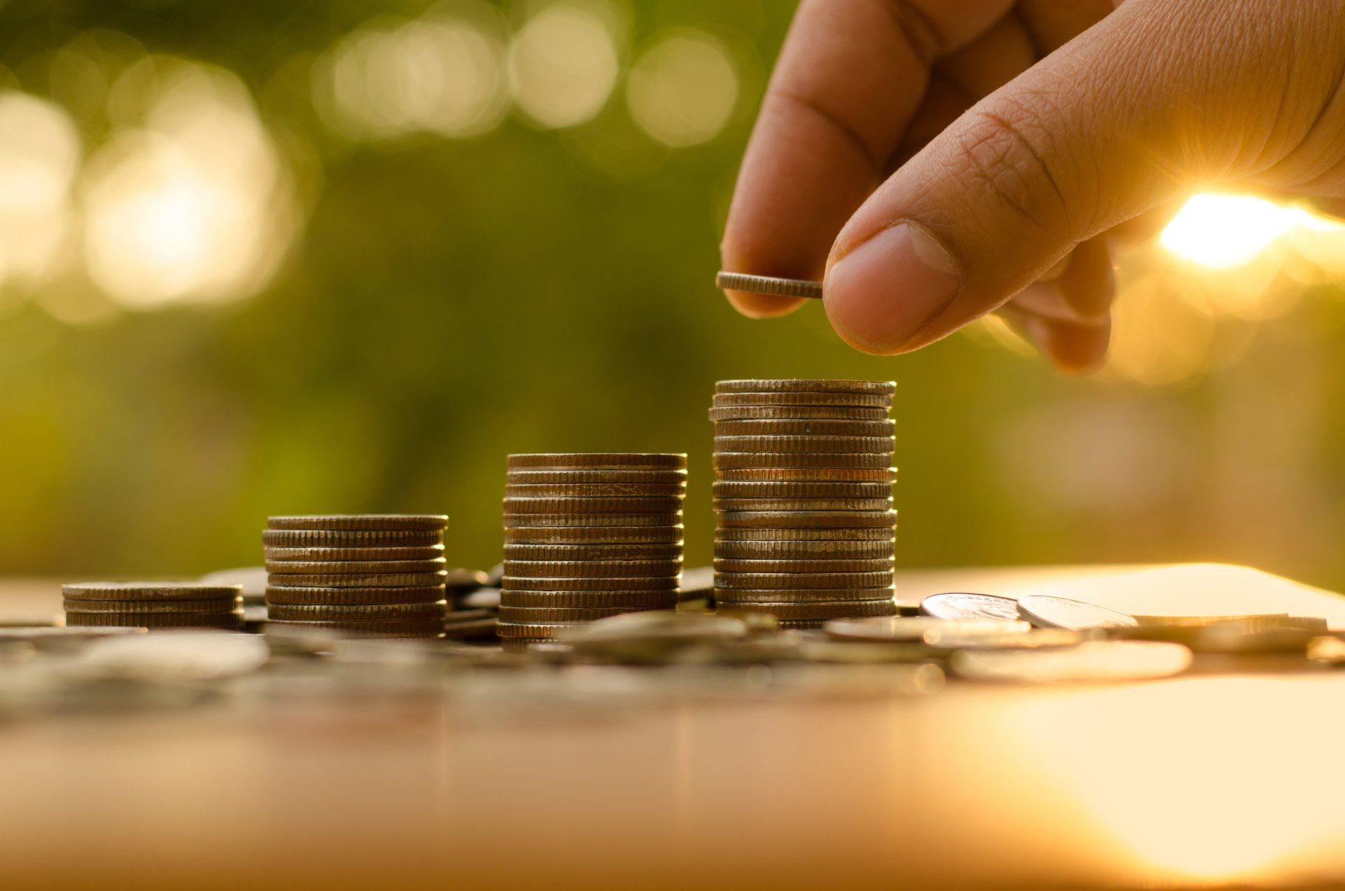 Hand adding coin to a stack of coins, symbolizing growth, with blurred green background.