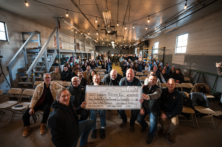 Group holds large check in front of a crowd, likely for a donation. Inside a large building with string lights.