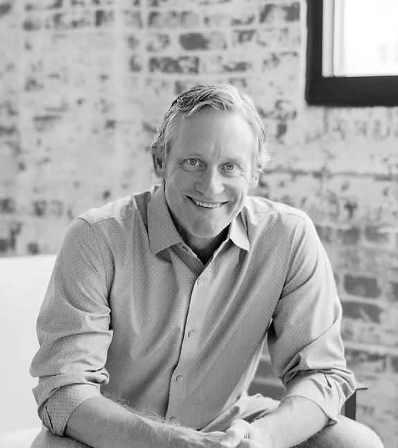 Man smiling, seated against a brick wall, wearing a button-down shirt.