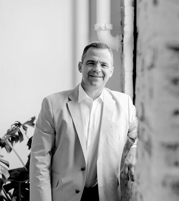 Man in a blazer smiles, leaning against a brick wall near a plant, outdoors.