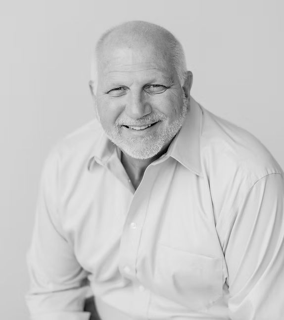 Smiling, balding man with a beard wearing a button-down shirt; waist-up shot against a plain background.