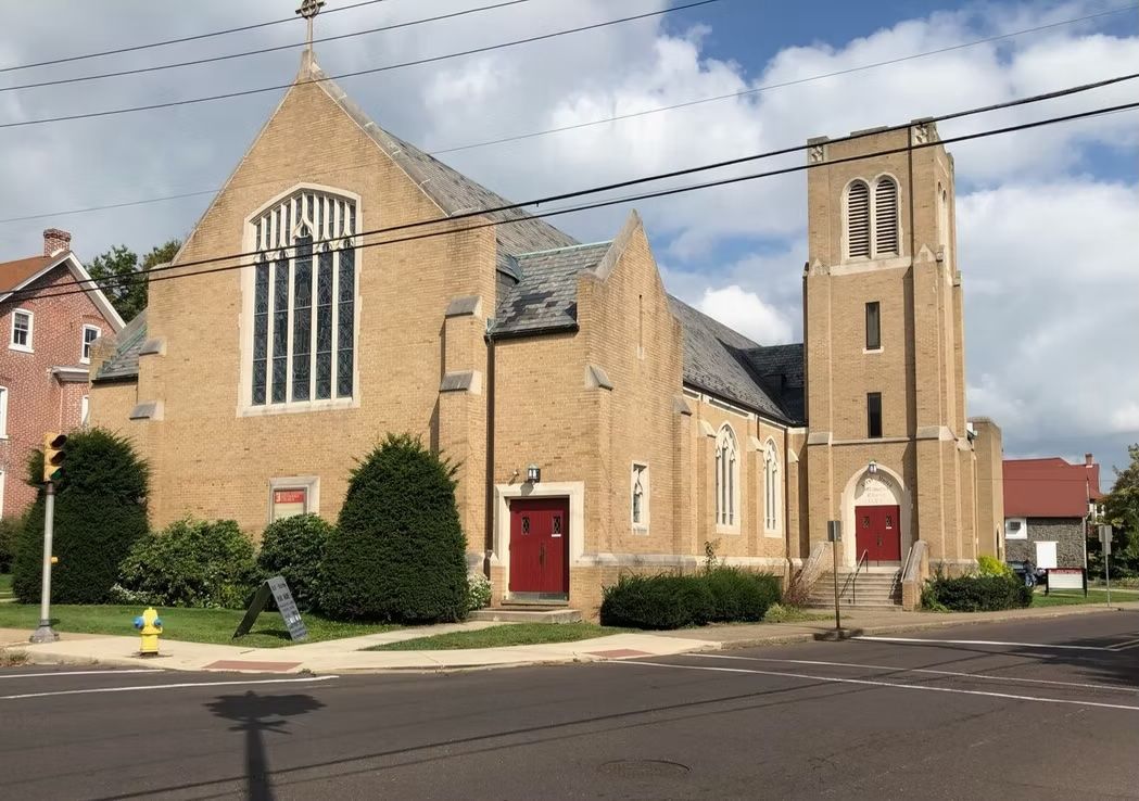 Brick church with steeple on a sunny day; red doors, green bushes, and utility wires.