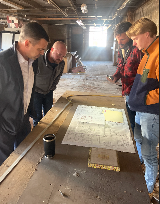 Four men examine a blueprint on a table in a dilapidated building. Sunlight streams through a window.