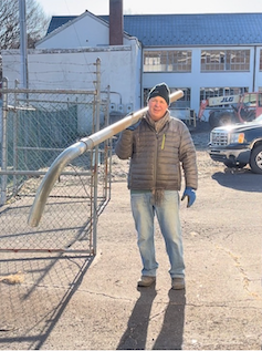 Man outdoors holding large metal pipe over his shoulder, smiling, wearing winter clothes.