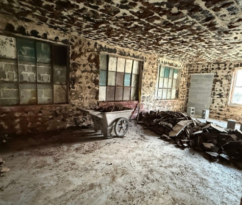 Dilapidated room with stone walls, industrial windows, and debris, wheelbarrow in center.
