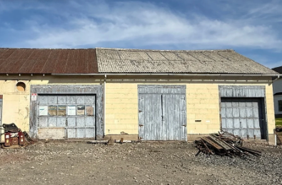 Yellow, weathered building with three gray garage doors, rusty roof. Debris in front, against blue sky.