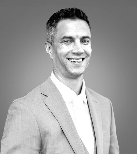 Smiling man in light gray suit, white shirt, against a gray background.