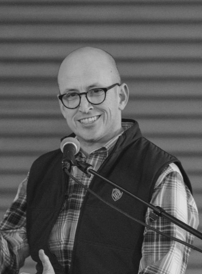 Bald man with glasses smiles into a microphone, wearing a vest and plaid shirt, speaking.
