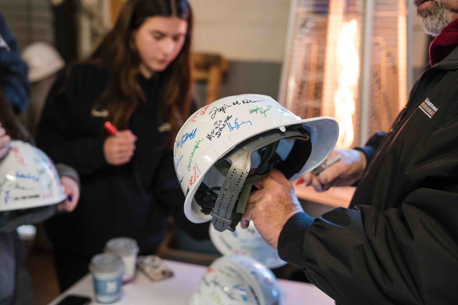 People sign white hard hats indoors, likely at a construction site celebration.