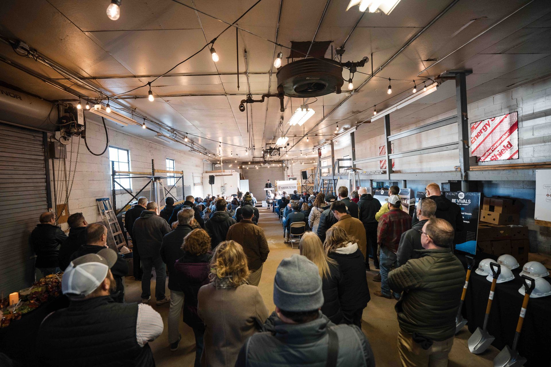 Large crowd in an open, industrial-style space, attending an event. People facing a central area, with string lights overhead.