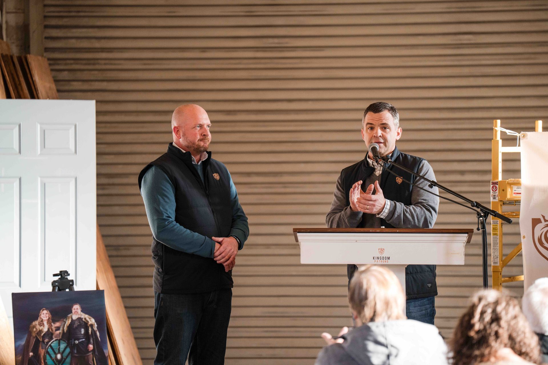 Two men at a podium, one speaking. Audience applauds in a warehouse setting.