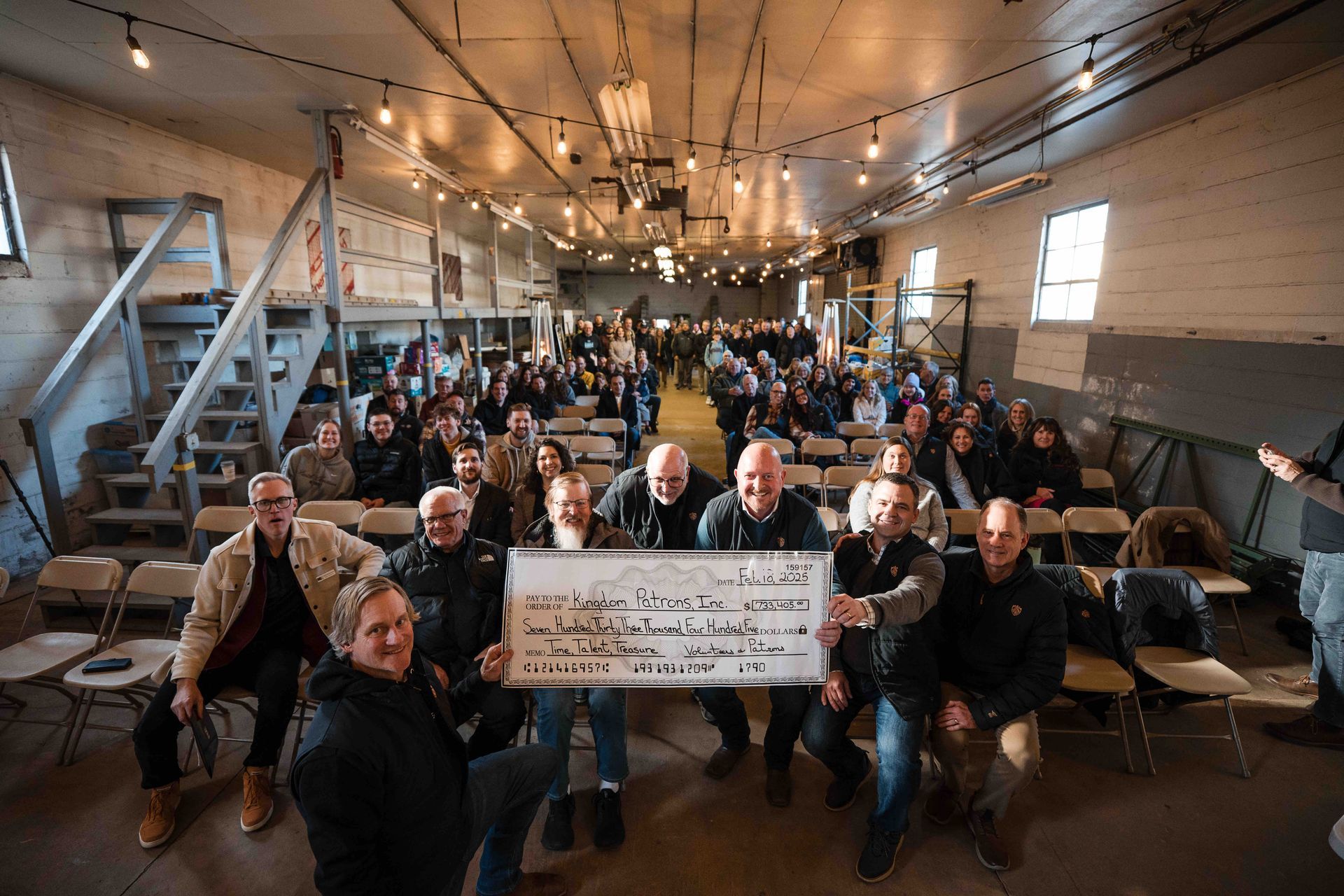 Large group in a rustic barn with a sign, celebrating, some smiling, lit by strings of lights.