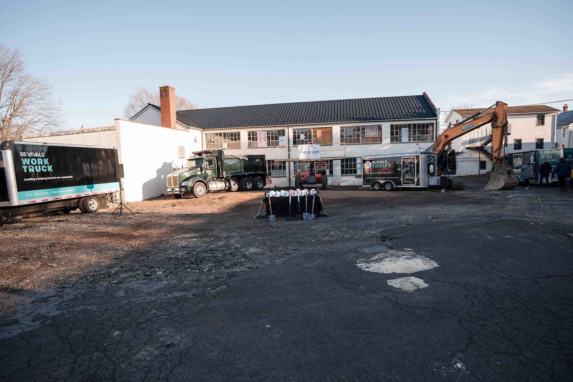 Demolition site. A two-story building is surrounded by construction vehicles. A group of people stand in front.