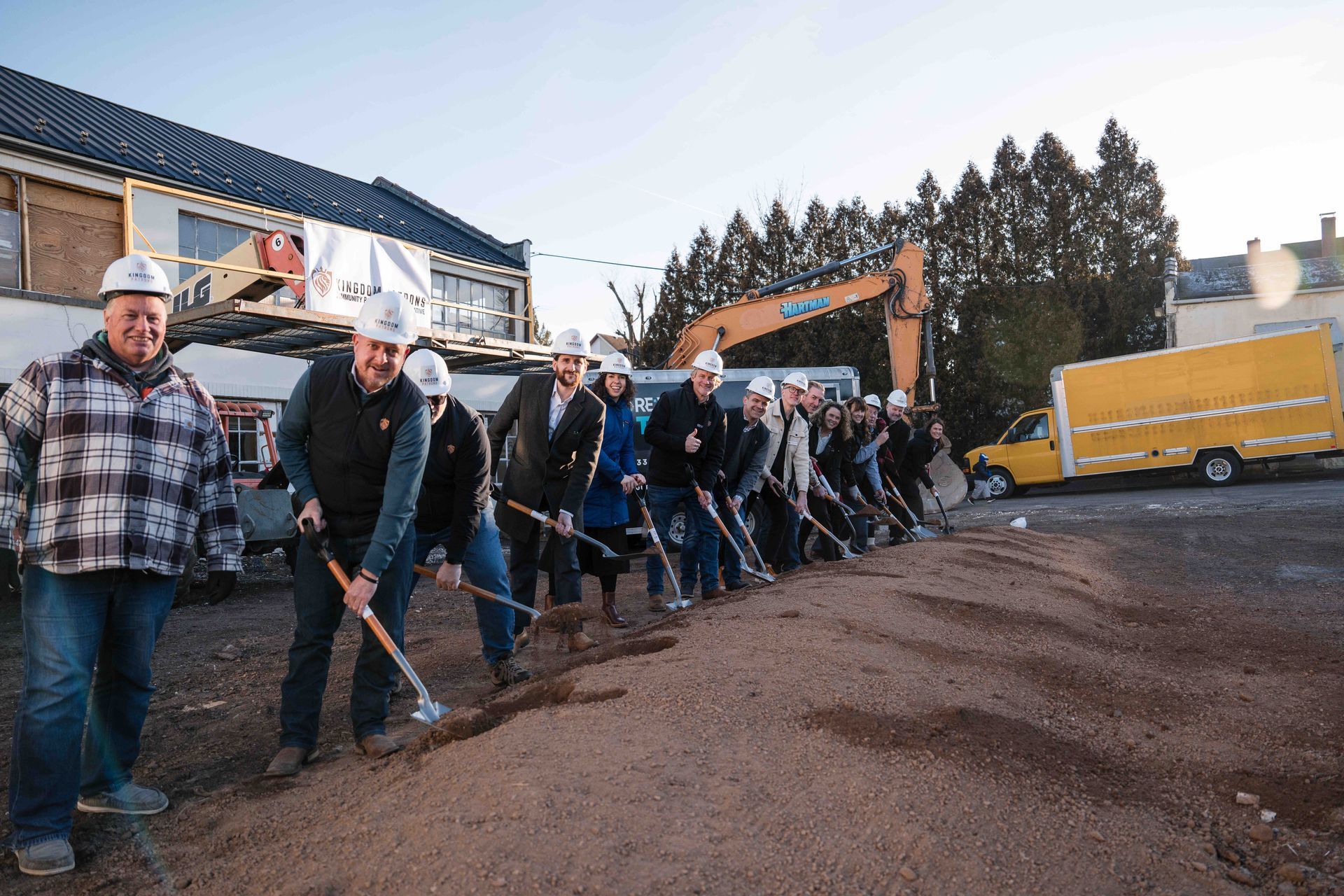 Group of people with shovels breaking ground at a construction site; a yellow truck is in the background.