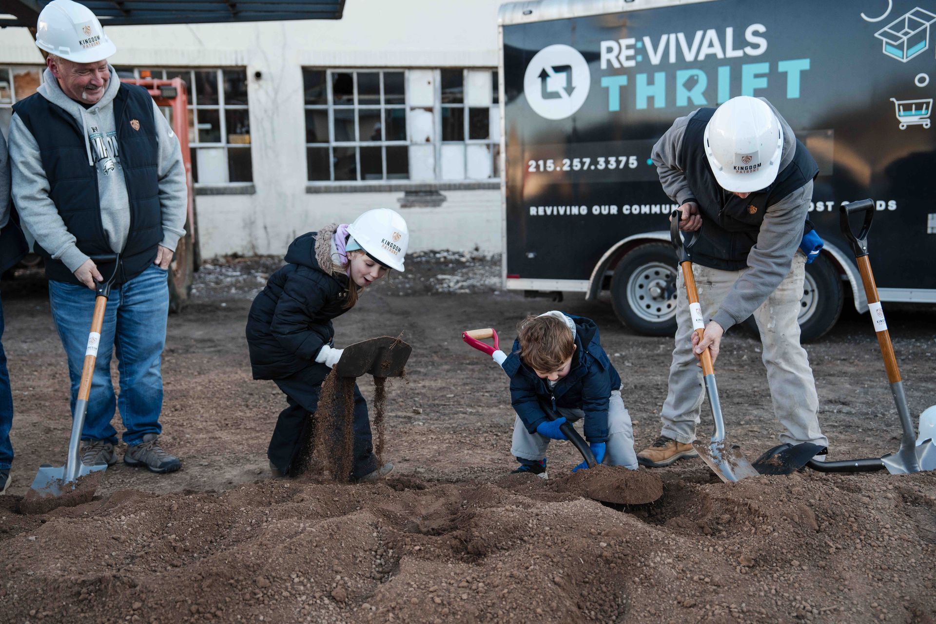 People in hard hats breaking ground for a thrift store; one child digs with a small shovel.