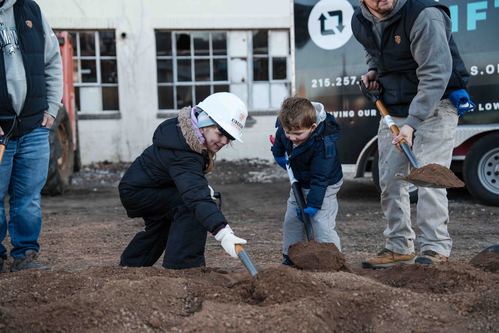Two children and a man shoveling dirt at a groundbreaking ceremony; white hard hat, gray sky.