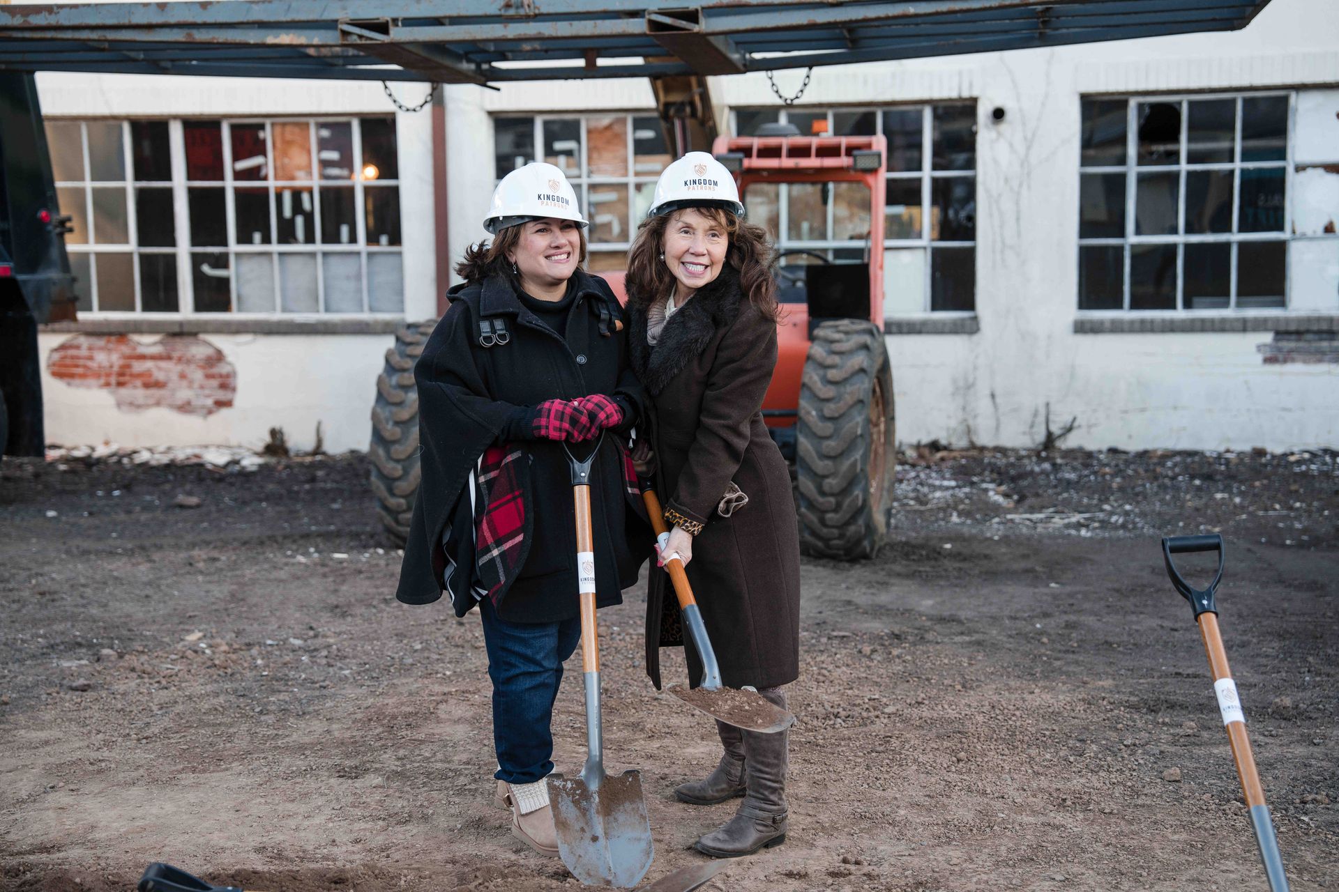 Two women in hard hats holding shovels at a ground-breaking ceremony; in front of a white building.