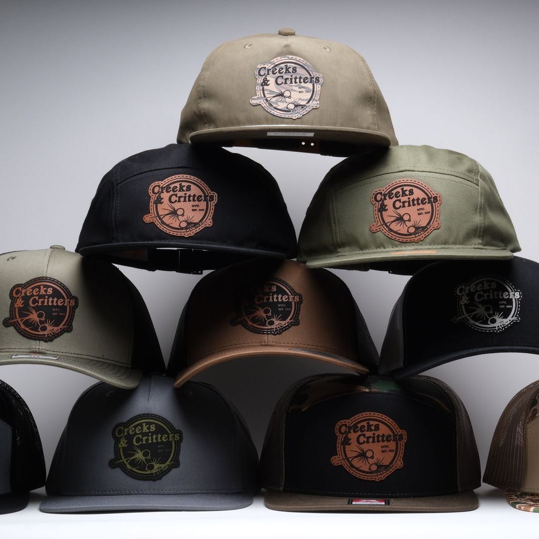 A bunch of hats are stacked on top of each other featuring custom leather patches for a fly fishing company