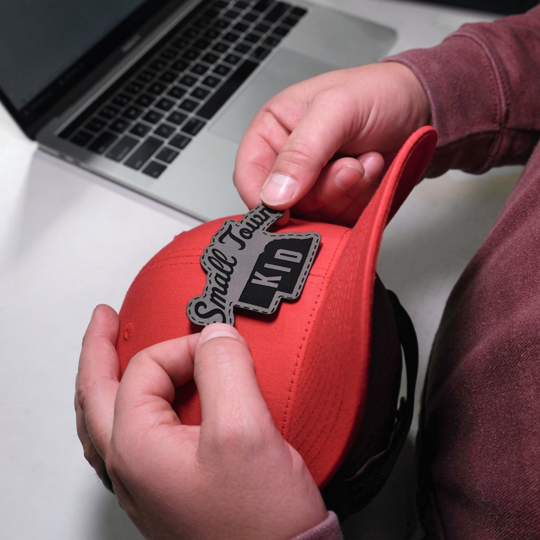 A person is putting a leather patch on a red hat that says small town kid