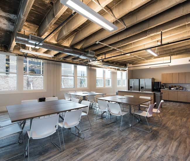 A modern office break room with tables, chairs, and a kitchen area, bright with natural light from windows.