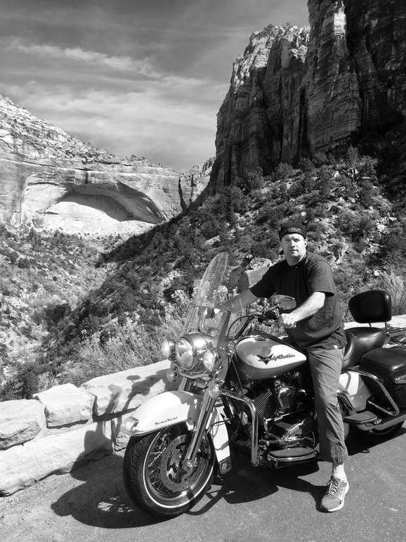 Man on a motorcycle poses on a scenic road with rock formations in the background.