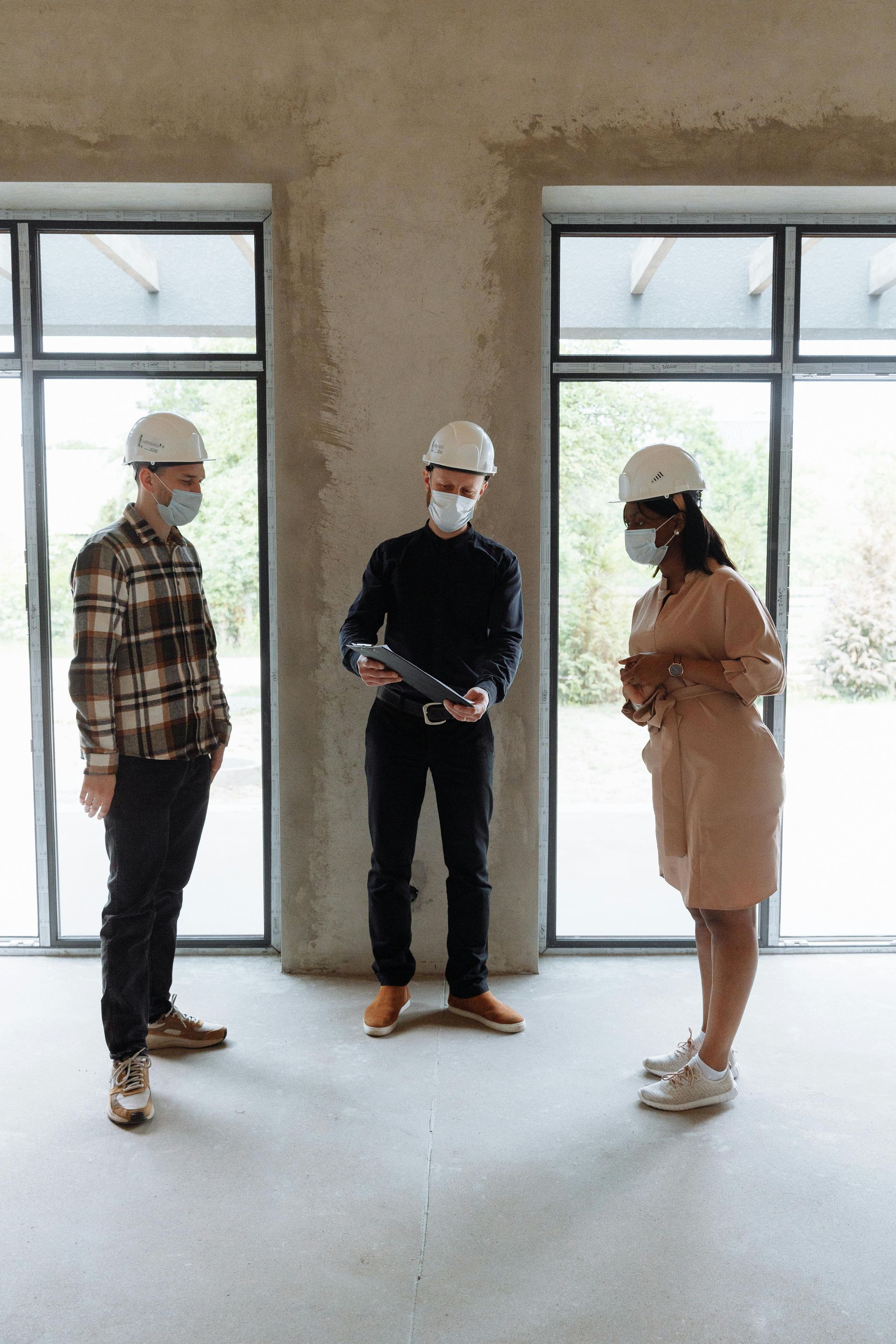 Three people in hard hats and masks discuss plans in a building under construction.