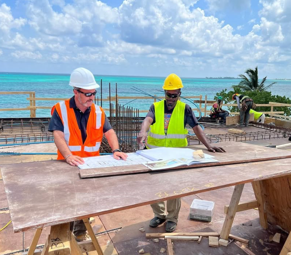 Two construction workers reviewing blueprints at a site near the ocean. Both wear vests and hard hats.