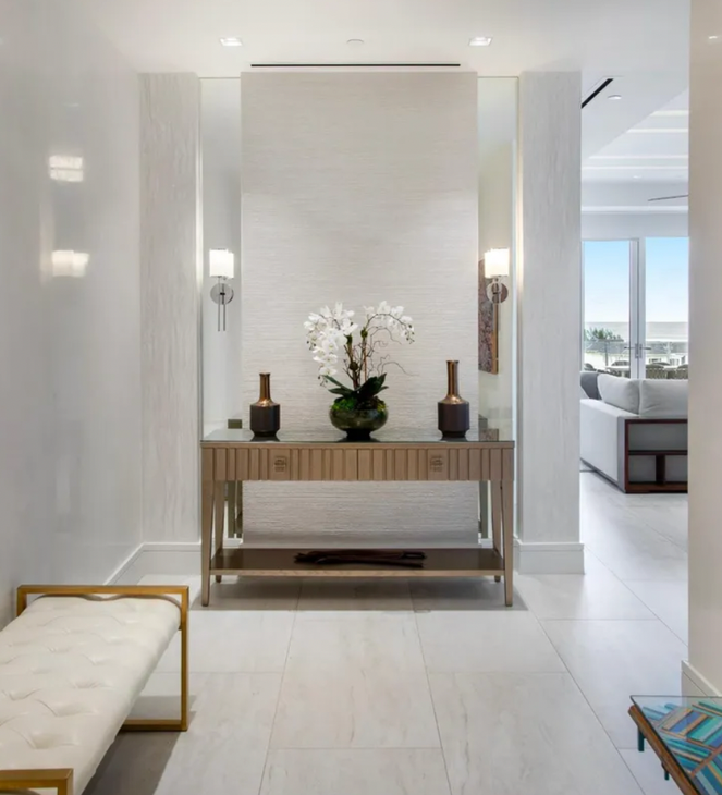 A bright hallway with a wooden table, vases, and orchids. White walls and flooring.