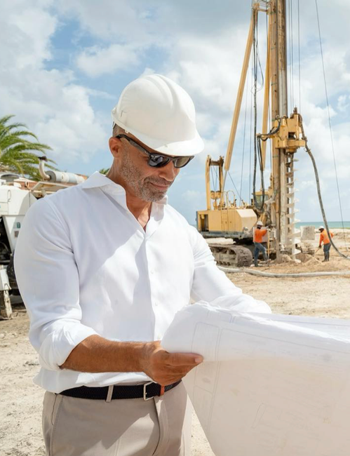 Construction worker in a white hardhat and sunglasses reviews blueprints on a construction site.
