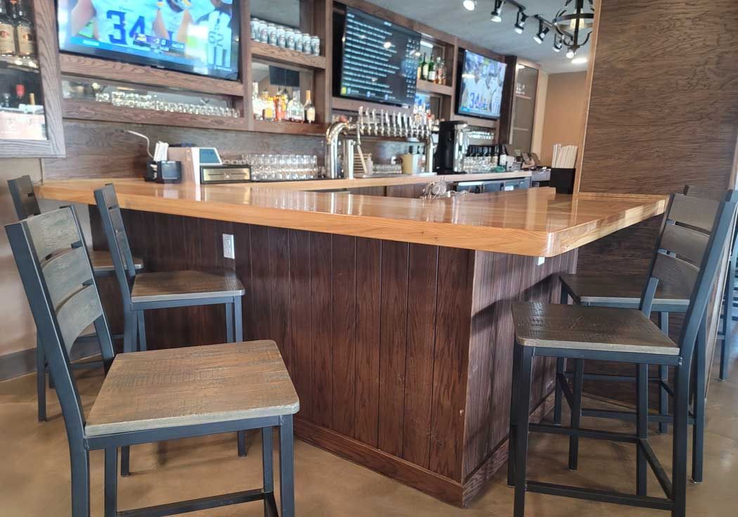 Bar with wooden counter, stools, and TVs. Light brown wood bar front, brown stools, multiple TVs above the bar.