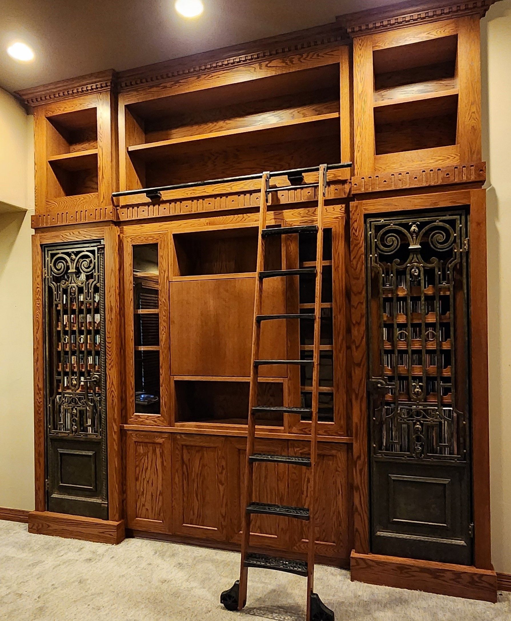 Wooden built-in bookcase with a rolling ladder. Two ornate dark doors flank glass-front cabinets. Beige walls and a light carpet.