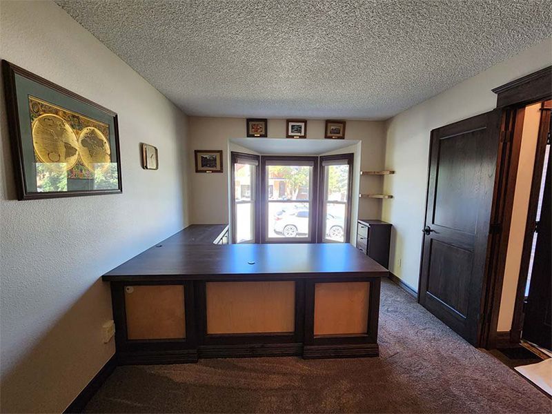 A home office with a large dark wood desk, bay window, and framed artwork on the walls.
