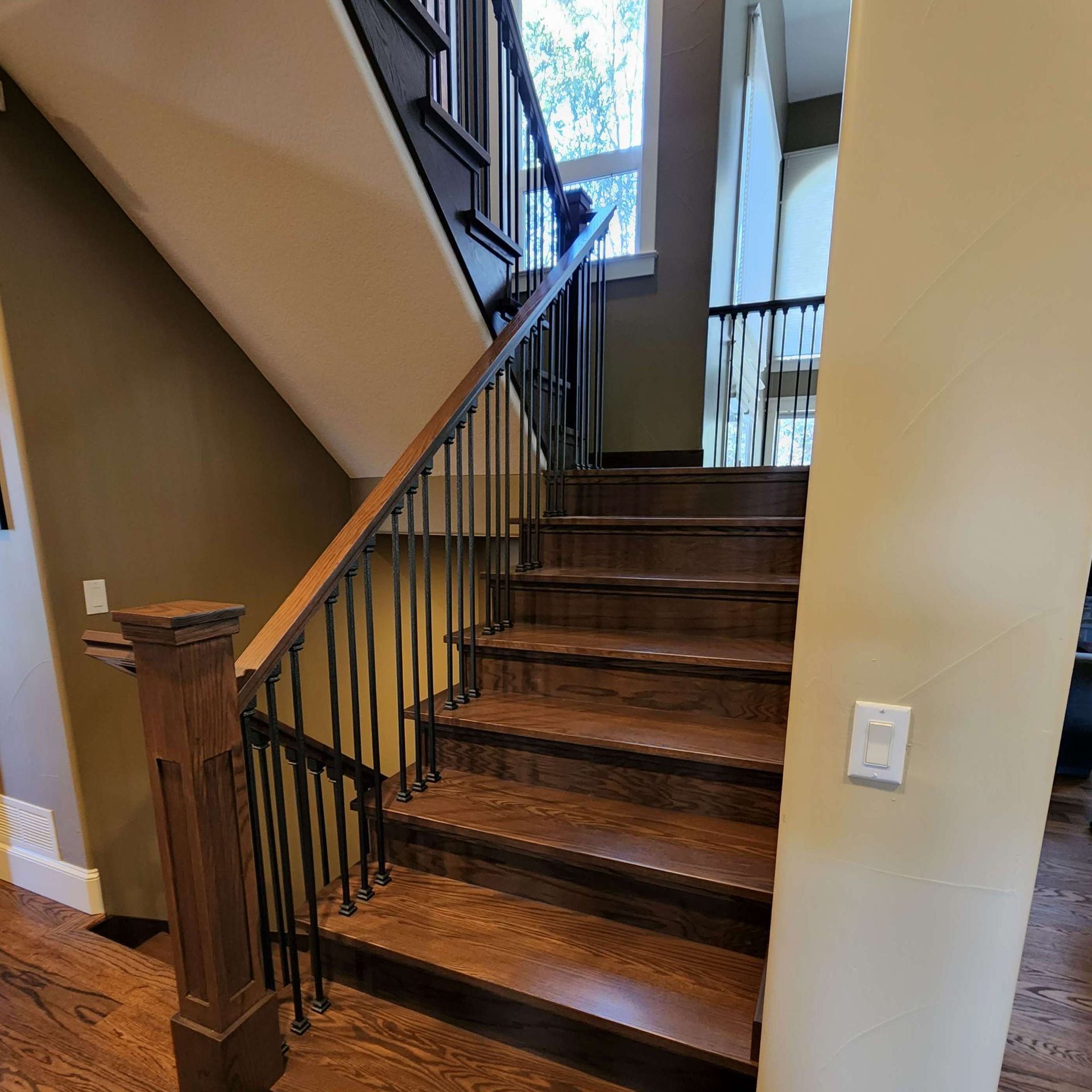 Staircase with wooden steps and railing, black iron balusters, tan walls, and a window at the top.