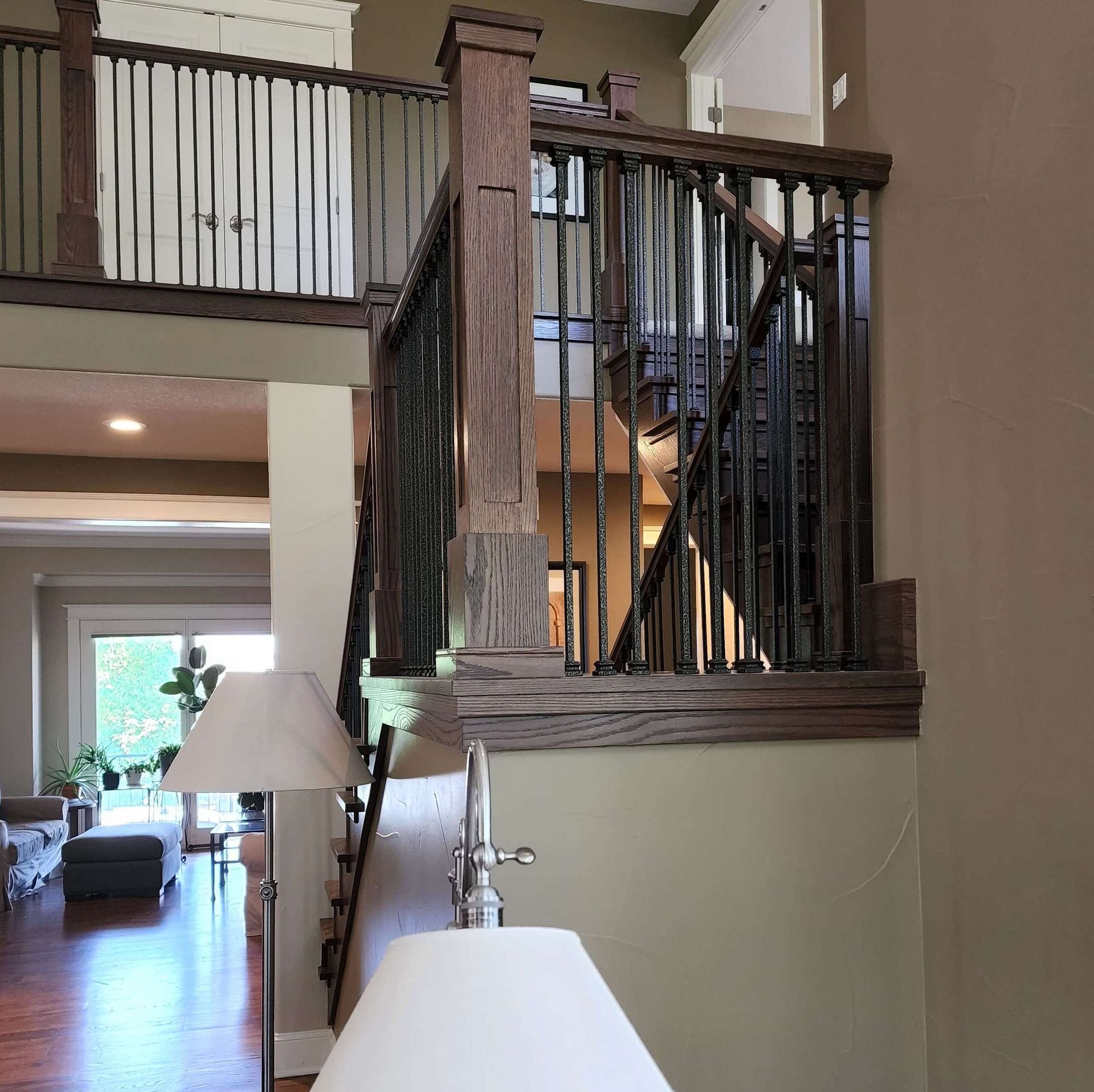 Staircase with wooden railing and wrought iron spindles. Beige walls, hardwood floors, and a lamp in the foreground.
