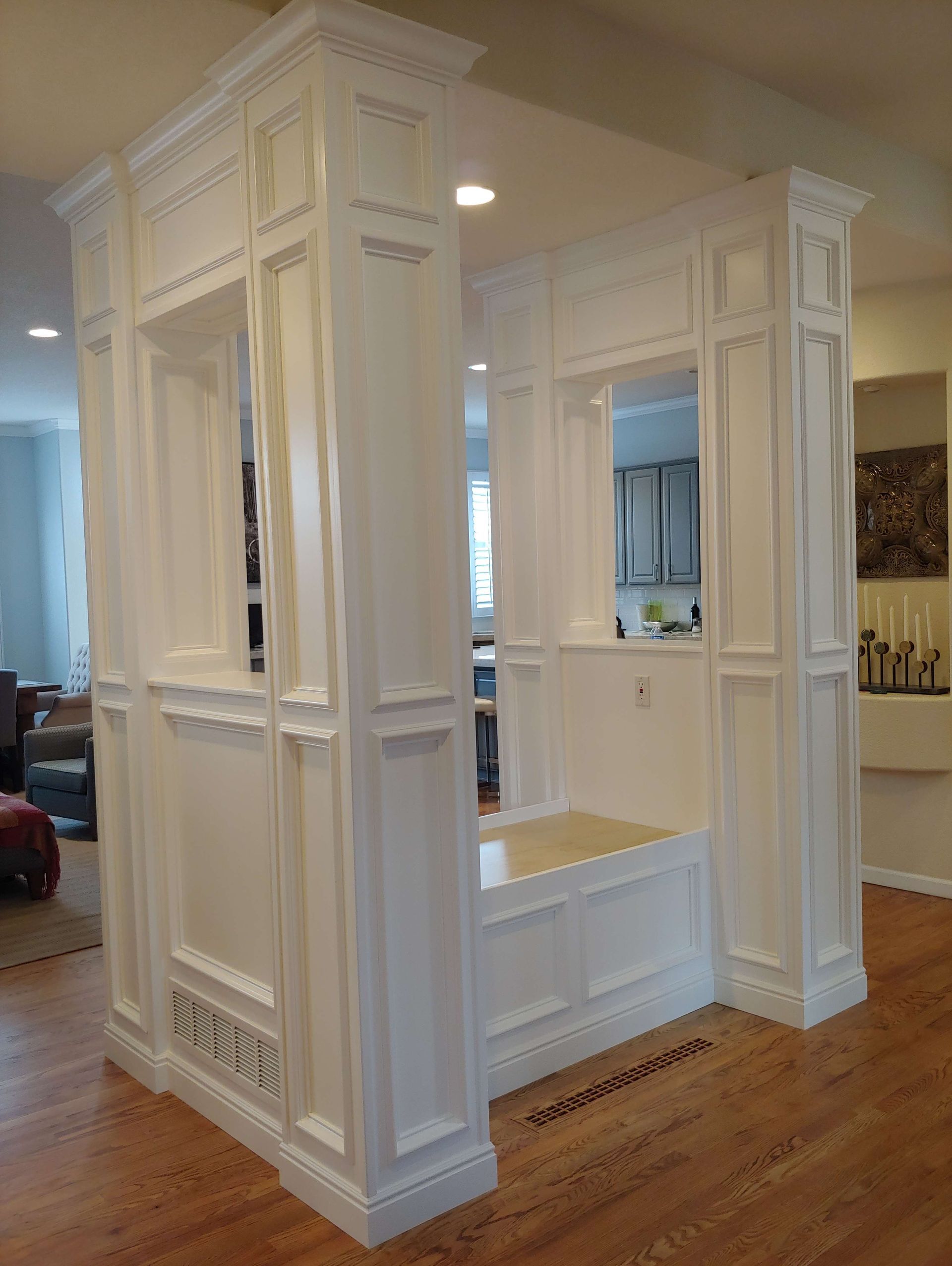 White built-in architectural structure with paneled walls, pillars, and a bench; in a room with hardwood floors.